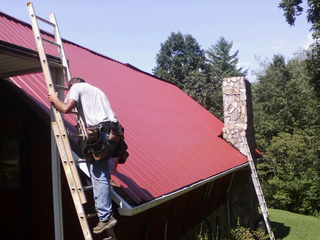 Roofer on a ladder beside a red metal roof and stone chimney — Soffit & Fascia Repair by Roofing Brothers in Ottertail